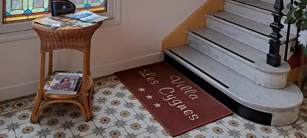 L'entrée de l'hôtel : tapis « Villa Les Cygnes », guéridon en rotin et premières marches de l'escalier