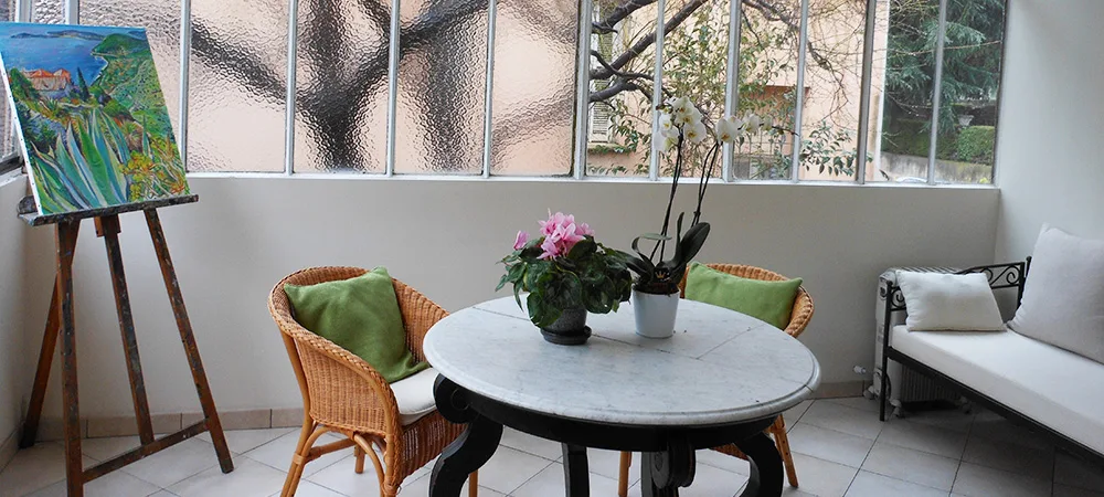 Salle du petit-déjeuner de l'hôtel : table de marbre, chaises en rotin et chevalet devant les vitres